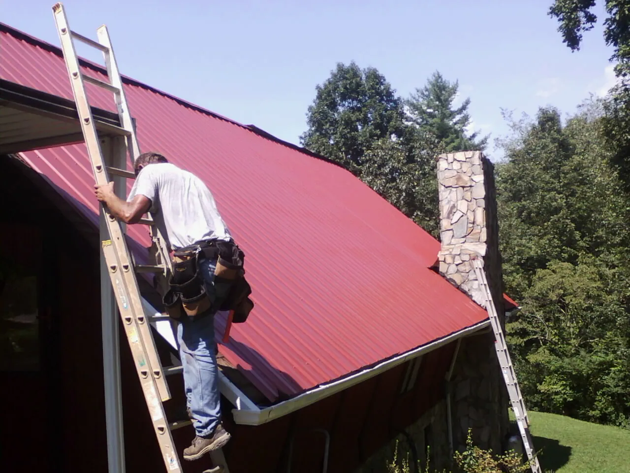 Roofer on a ladder beside a red metal roof and stone chimney — Soffit & Fascia Repair by Roofing Brothers in Elkhart